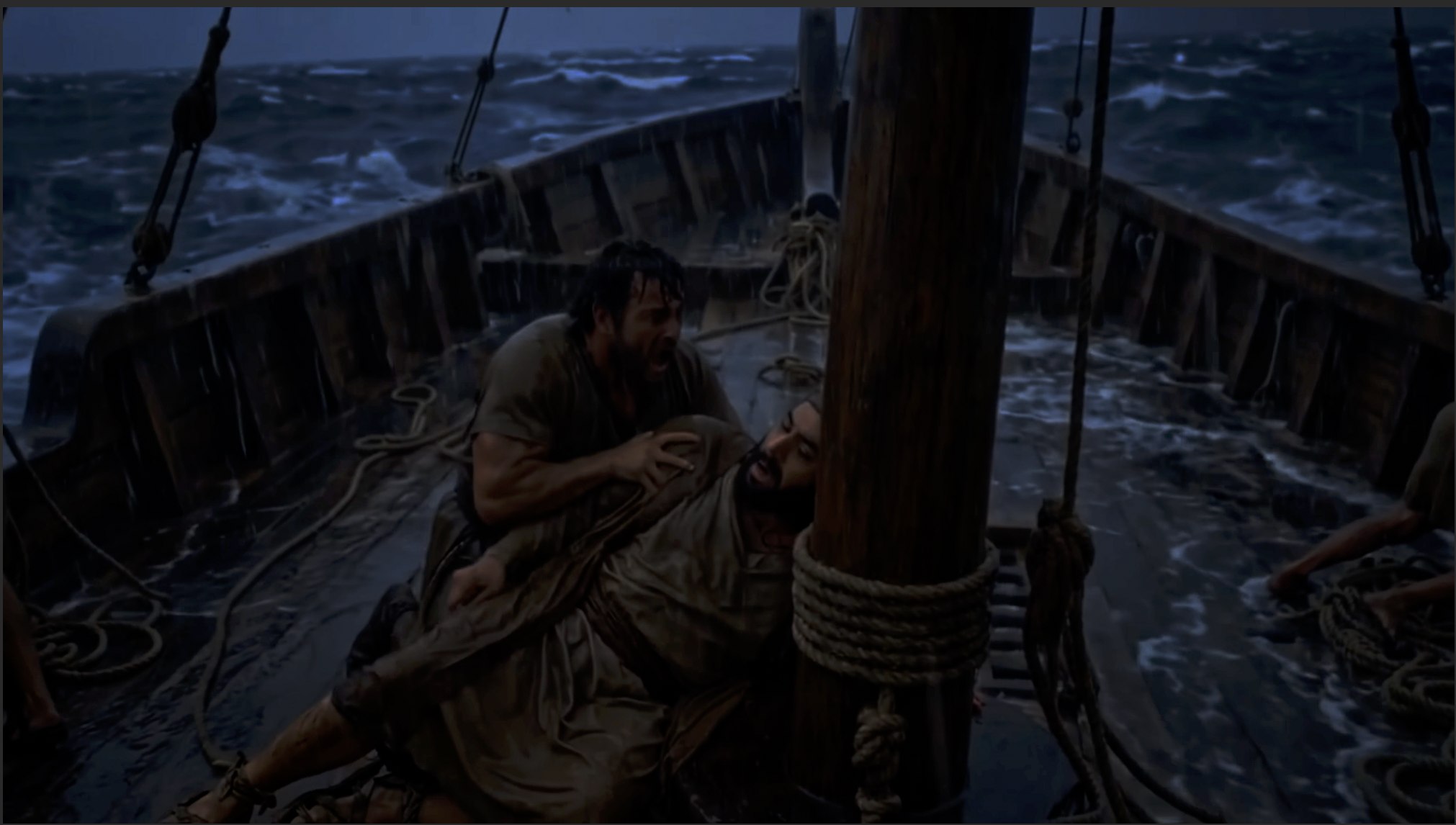 Two men struggle on the deck of a storm-tossed ship in the dark, waves crashing around them